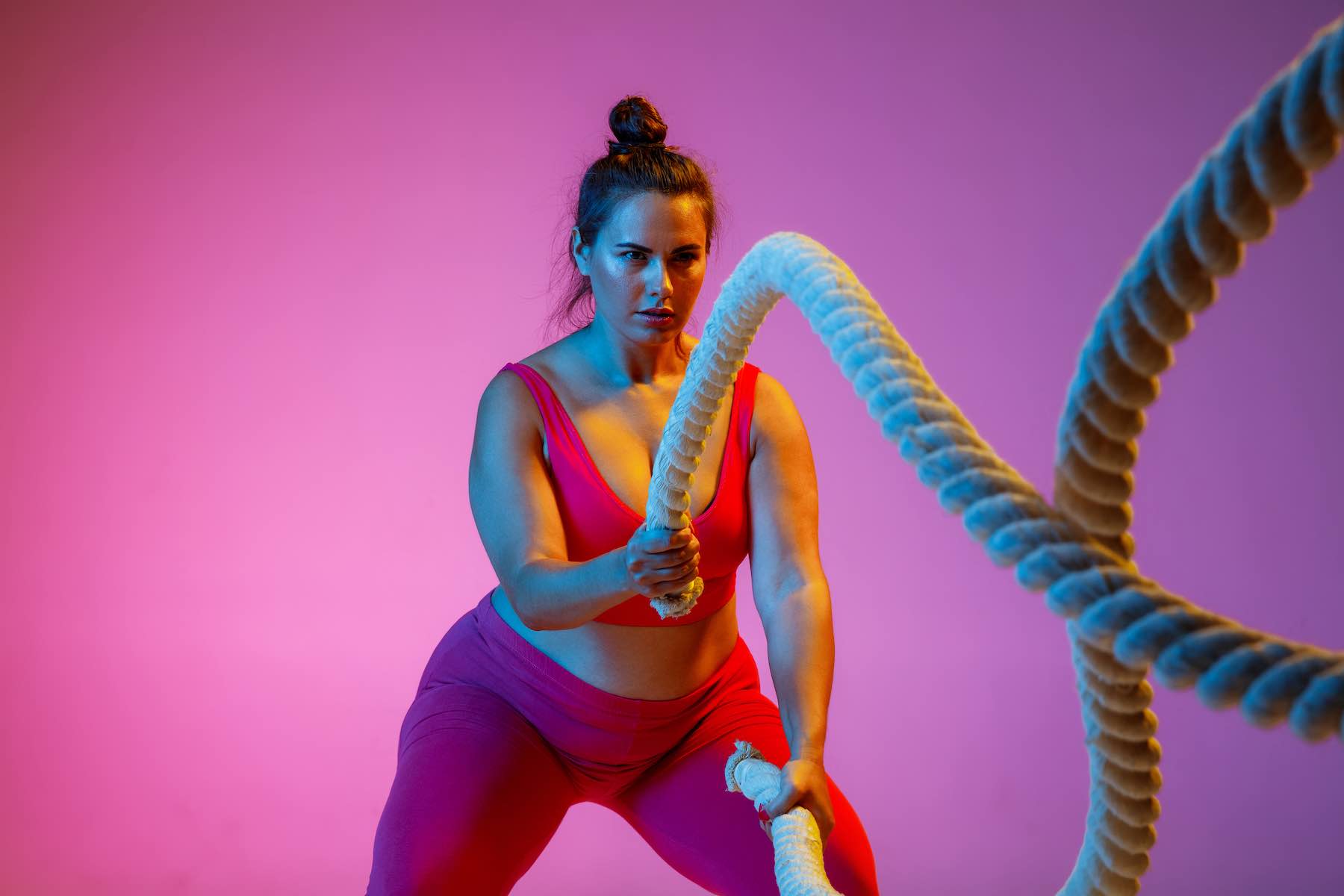 Young caucasian plus size female model's doing exercises on gradient purple background in neon light. training with the rope. Concept of sport, healthy lifestyle, body positive, equality.