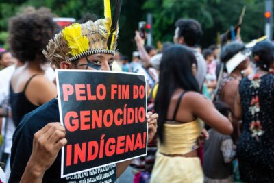 Homem indígena com cocar protestando com faixa "pelo fim do genocídio indígena", em marcha pelos direitos dos povos indígenas.